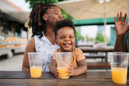 Portrait of a happy little boy holding an orange juice in his hands and looking at the camera while sitting in an outdoor coffee shop with his parents