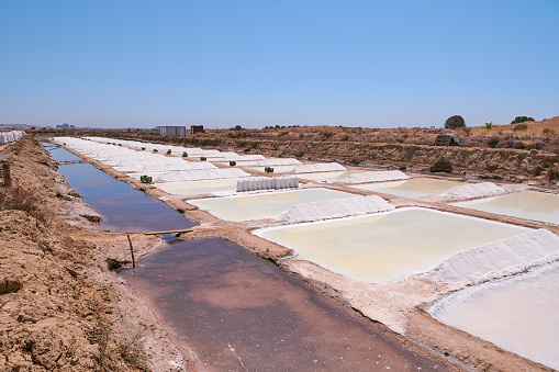 Castro Marim traditional Salt Flats on a summer sunny day. Sea salt is piled between the ponds, and a few is already packed in white bags.  The perspective creates a vanishing point to the left. These salt flats are located in the south of Portugal, by the Guadiana River, near the Spanish border. The area is named Sapal de Castro Marim and Vila Real de Santo António Natural Reserve.