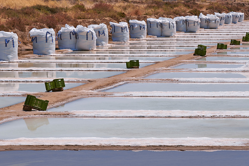 Detail of a salt flat, with some plastic green boxes and white big bags of salt. The ponds and the reflection from the sun, form a geometrical pattern. Photo taken at Castro Marim traditional Salt Flats on a summer sunny day. These salt flats are located in the south of Portugal, by the Guadiana River, near the Spanish border. The area is named Sapal de Castro Marim and Vila Real de Santo António Natural Reserve.