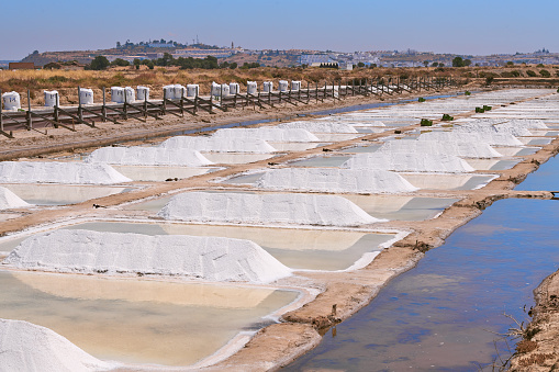 General view of salt flats in harvesting time, with piles of sea salt, and white big bags in the background. Photo taken at Castro Marim traditional Salt Flats on a summer sunny day. These salt flats are located in the south of Portugal, by the Guadiana River, near the Spanish border. The area is named Sapal de Castro Marim and Vila Real de Santo António Natural Reserve.