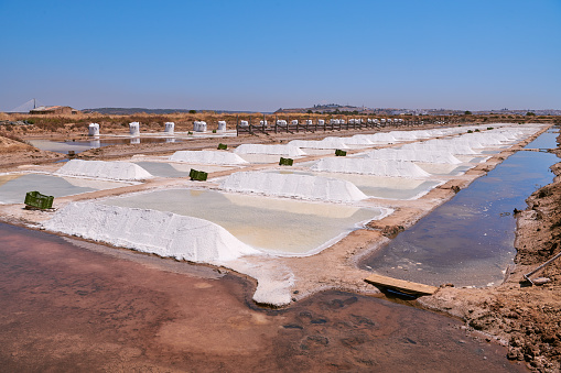 General view of a traditional sea salt exploration, in Castro Marim. Photo taken  on a summer sunny day, in harvest time. Salt is piled between the ponds and white big bags, full of salt can be seen on the background. The perspective creates a vanishing point to the right.  These salt flats are located in the south of Portugal, by the Guadiana River, near the Spanish border. The area is named Sapal de Castro Marim and Vila Real de Santo António Natural Reserve.
