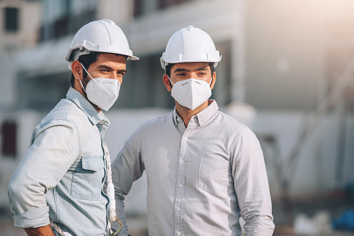 Two Caucasian man waering protective mask working in construction site, engineers and architects working together at real estate work site project in covid 19 pandemic situation.