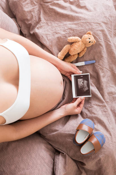 Pregnant Woman sitting on bed and Looking At Ultrasound Scan photo of her Baby. A pregnant female shows her ultrasound report and her unborn baby stock photo