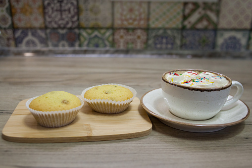 Two muffins with raisins on a wooden tray and a cup of hot chocolate with marshmallows and colored sprinkles.