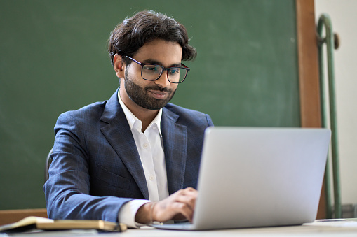 Young indian business man executive wearing suit working on laptop sitting in office at work desk. Arabic manager worker using software on computer technology checking digital professional data.