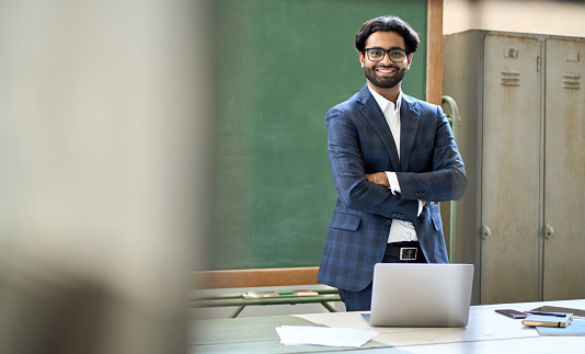 Happy young adult indian business man coach wearing suit looking at camera standing in office. Arab college teacher or university professor posing for portrait at workplace desk in classroom.