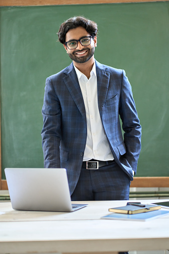 Smiling young indian business man manager wearing suit looking at camera standing in office. Arab teacher or professor posing for portrait at work desk with laptop in front of blackboard. Vertical
