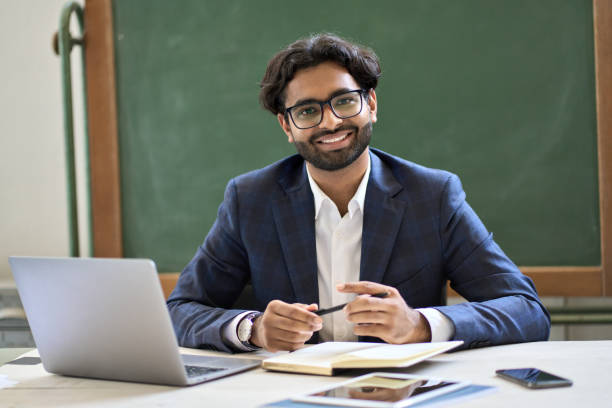 Happy young indian businessman teacher sitting at work in classroom. Portrait Happy young indian arabic businessman professional coach, teacher or university professor wearing suit looking at camera sitting at work desk in classroom office posing for portrait at workplace. professor business classroom computer stock pictures, royalty-free photos & images