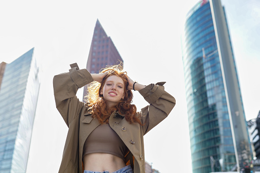 Happy teen smiling stylish redhead girl standing on big city urban street skyscrapers background looking at camera, enjoying freedom, feeling joyful and motivated in downtown outdoor.