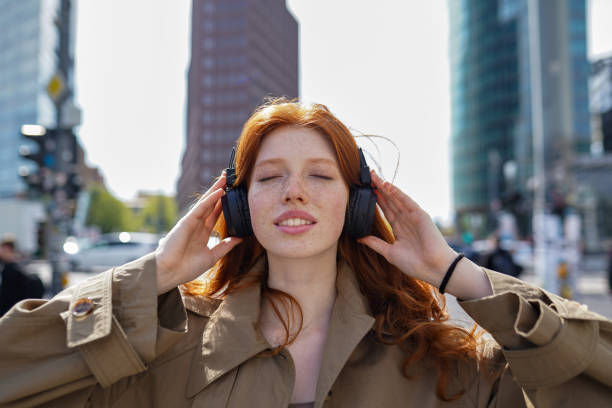 счастливый подросток рыжеволосая девочка в наушниках слушает музыку в большом городе. - gen z стоковые фото и изображения