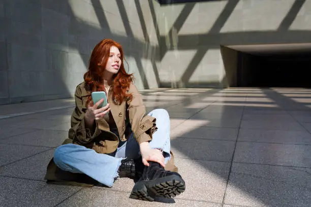 Redhead cool teenage hipster fashion girl user using smartphone holding phone checking social media posts on cellphone sitting on urban city walls background lit with sun light.