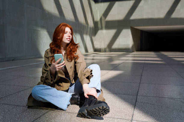 рыжеволосая крутая девочка-подросток с помощью телефона сидит на фоне городских стен. - gen z стоковые фото и изображения