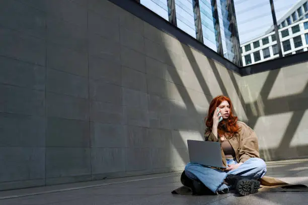 Teen redhead hipster girl student talking on cell phone using laptop computer modern technology connected devices in city urban location outdoors.