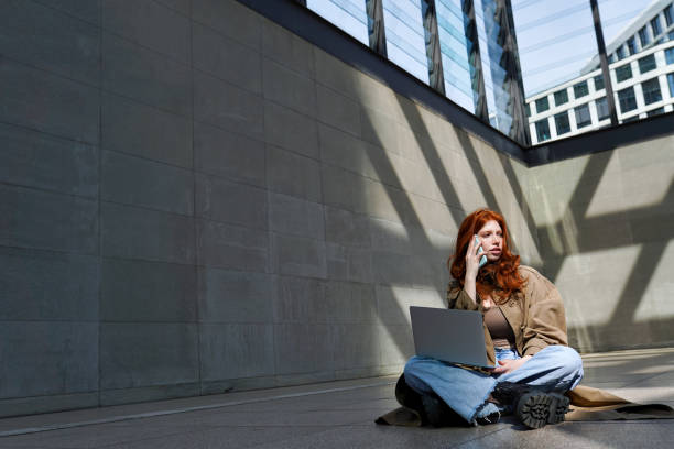 подросток рыжеволосая девочка с помощью ноутбука разговаривает по сотовой связи в городском городе. - gen z стоковые фото и изображения