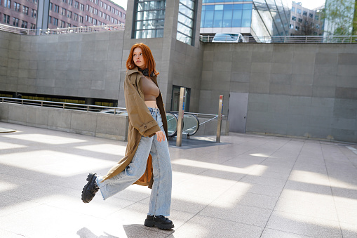 Cool teen stylish redhead fashion hipster girl model walking in big city subway. Beautiful teenage girl with red hair wearing trench coat going in urban location.