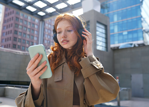 Teen redhead girl standing on urban street skyscrapers background wearing headphones using smartphone listening to music, audio book, travel guide or podcast while walking in big city.