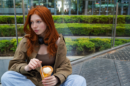 Cool teenage hipster girl eating ice cream on big city urban street. Pretty teen generation z girl with red hair wearing trench coat enjoying icecream spending time in park outdoors.