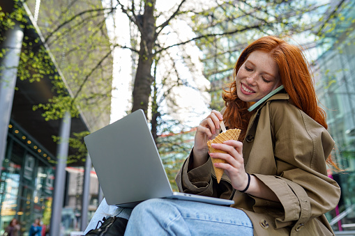 Happy teen redhead hipster girl student using laptop, taking on the phone while eating ice cream in city, online learning outdoors, sitting on urban street spending time outside.