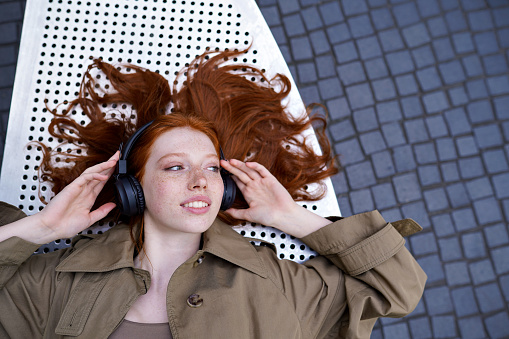 Teen redhead hipster girl wearing headphones listening to mobile streaming music mix, audio book, travel guide or podcast enjoying modern sound tech outdoors looking at copy space. Top view