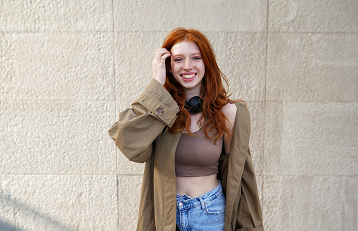 Happy teen stylish cool redhead fashion girl model standing on urban wall background looking at camera. Portrait of beautiful smiling teenage girl with red hair wearing trendy clothes laughing outdoor