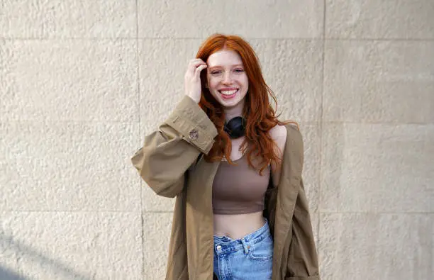 Photo of Happy teen redhead girl looking at camera standing on urban wall background.