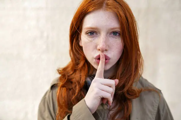 Photo of Hipster teen redhead girl showing shh sign on urban wall background.