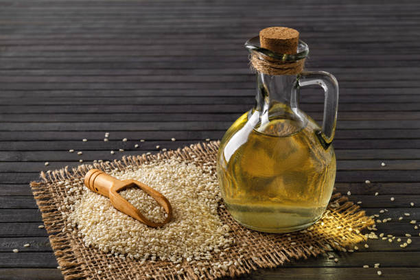 Fresh sesame oil in a glass jug and heap of white til seeds over black background. Vegetable oil from Sesamum indicum. Organic benne seeds for boosting immunity diet and calcium source. stock photo