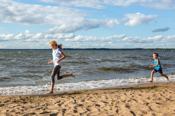 Side view of a girl running barefoot at the beach on a windy day. Her brother can be seen at the right side of the image, racing to catch up to his sister.