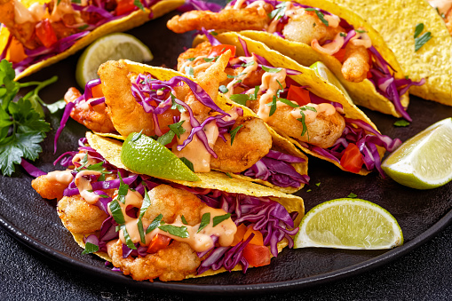 Bang Bang Shrimp Tacos with purple cabbage, tomatoes, parsley, lime drizzled with mayonnaise hot chili sauce on black plate on concrete table, horizontal view from above, close-up