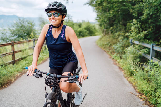 retrato de una mujer sonriente feliz vestida con ropa de ciclismo, casco y gafas de sol montando una bicicleta en el carril bici de asfalto fuera de la ciudad. imagen de concepto de personas deportistas activas. - cyclo cross fotografías e imágenes de stock