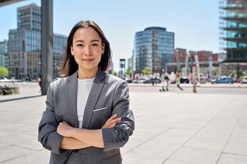 Young confident smiling Asian business woman standing on busy street, portrait. Proud successful female entrepreneur wearing suit posing with arms crossed looking at camera in big city outdoors.