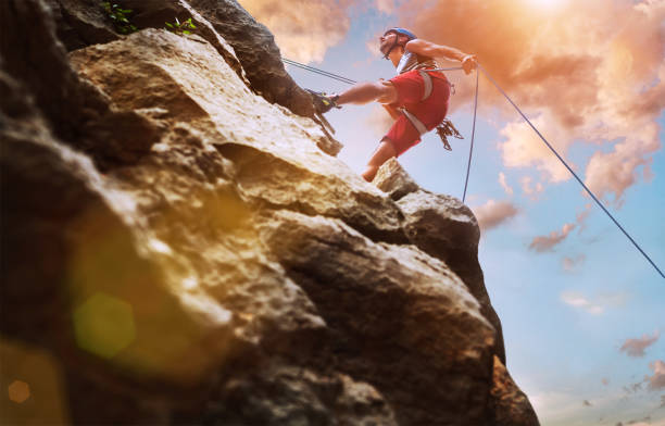 istock Muscular climber man in protective helmet abseiling from cliff rock wall using rope Belay device and climbing harness on evening sunset sky background. Active extreme sports time spending concept. 1414068959
