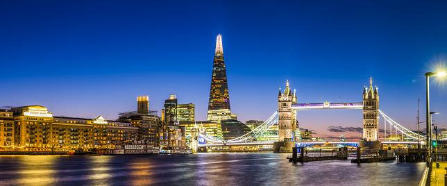 Panoramic view across the River Thames to the historic span of Tower Bridge illuminated against the blue dusk sky overlooked by the futuristic glass spire of The Shard in the heart of London, Britain's vibrant capital city.