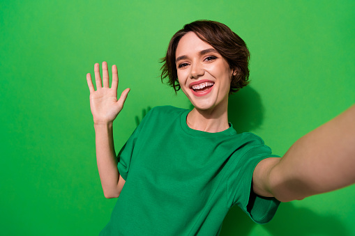 Photo of cute charming woman wear casual t-shirt recording self video waving arm palm isolated green color background.