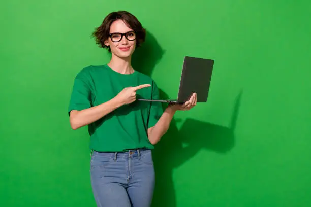 Photo of Photo of cute sweet woman wear casual t-shirt arm spectacles pointing modern device isolated green color background