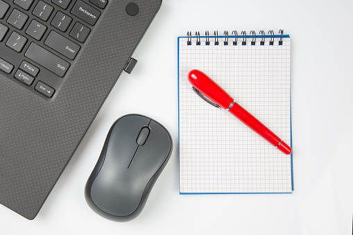 Computer wireless mouse, pen and notepad next to a laptop on a white table close-up. Items for office work