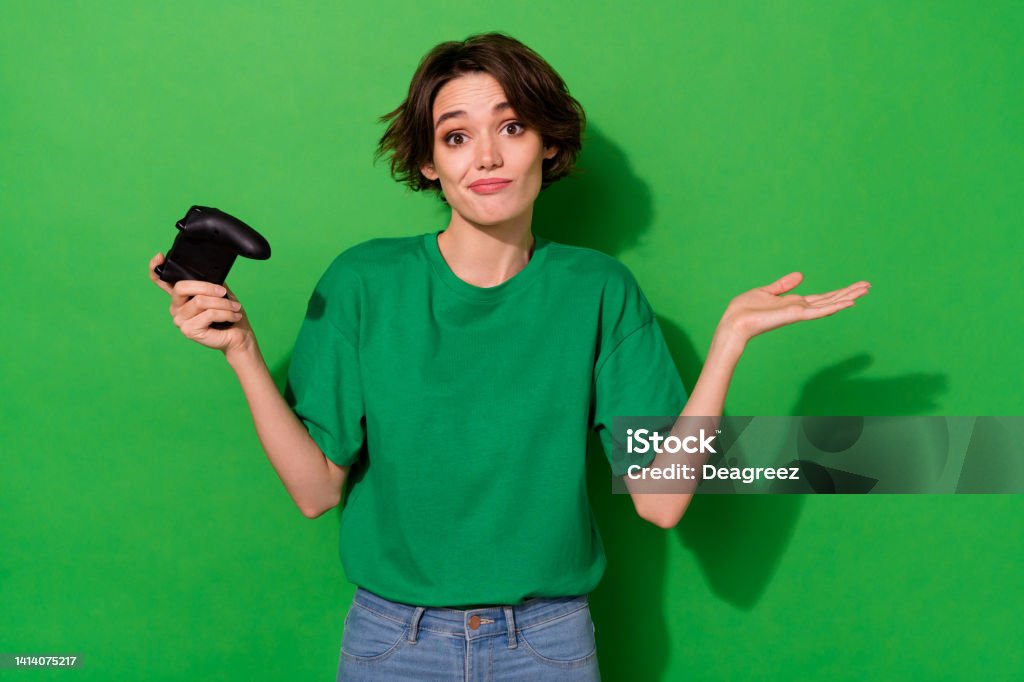 Photo of uncertain funny woman dressed summer t-shirt holding playstation shrugging shoulders isolated green color background Photo of uncertain funny woman dressed summer t-shirt holding playstation shrugging shoulders isolated green color background. Gamer Stock Photo