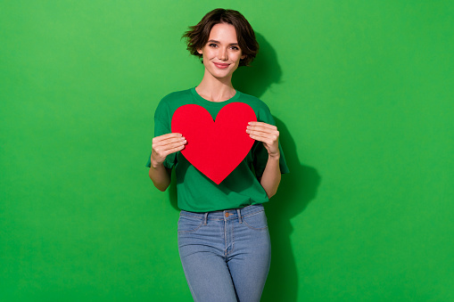 Photo of dreamy adorable woman dressed summer t-shirt holding red heart isolated green color background.