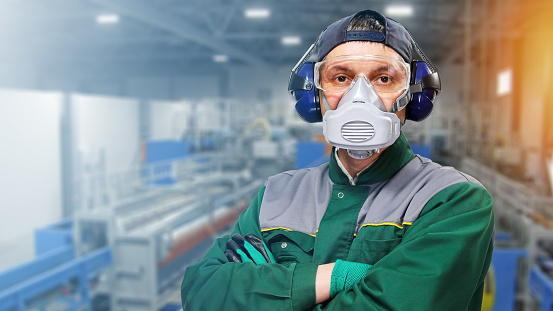 Worker in protective clothes, headphones and respirator stands against background of industrial factory, looks into camera