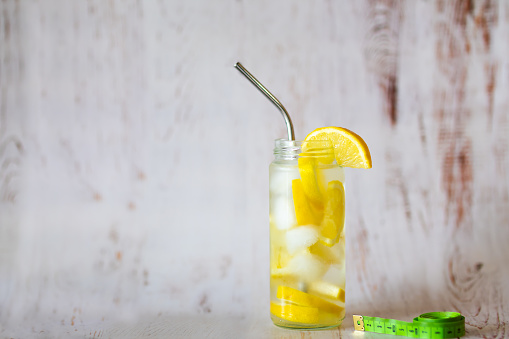 Lemon water in a glass jar with cut lemon and ice cubes and drinking straw, green measuring tape on the background. Loosing weight concept, detox