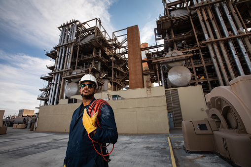 Portrait of an African-American man wearing coveralls, sunglasses, a hard hat and safety gloves with a coiled cable over one shoulder posing in front of a gas powered power station.
