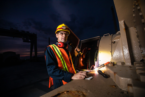 Portrait of a power plant operator wearing coveralls, had hat, high vis vest, and safety glasses working on a turbine at night.