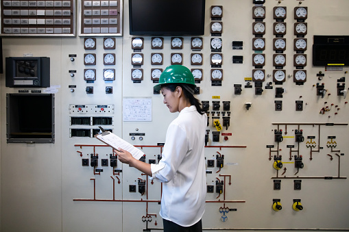 A young Chinese-American woman wearing a blouse and a hard hat working in the control room of a natural gas fired power plant.