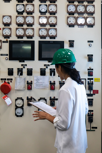 A young Chinese-American woman wearing a blouse and a hard hat working in the control room of a natural gas fired power plant.