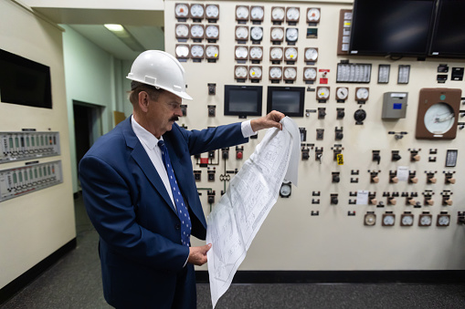 A man in his 70s  wearing a suit and a hard hat working in the control room of a natural gas fired power plant.