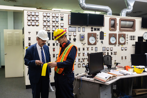 A man in his 70s wearing a suit and a hard hat and a younger technician wearing coveralls and safety gear working in the control room of a natural gas fired power plant.