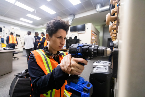 Non-binary mechanic wearing coveralls and safety gear using a drill to repair a panel in power plant the control room of a natural gas fired power plant.