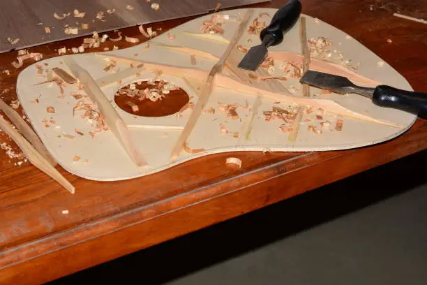 Selective focus on shavings and inside of Dreadnought Guitar, bracing, chisels. Shavings are created by voicing the soundboard of the Engelmann Spruce soundboard with cedar and Sitka Spruce bracing.