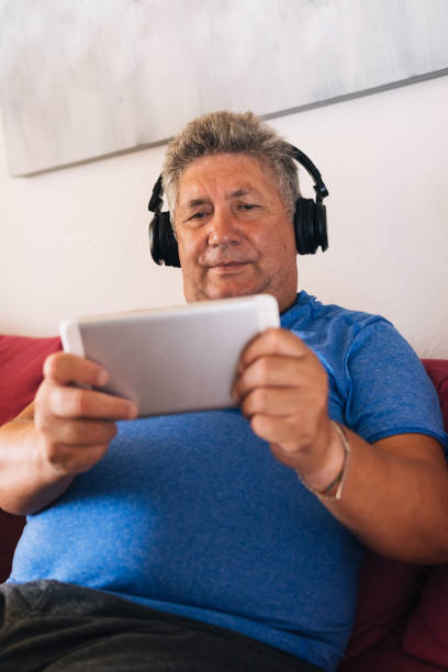 istock Retired white-haired man with black headset using his tablet to play online games. Vertical image of elderly person chatting on social networks while listening to music. Concept of technology 1414084778