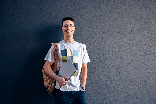 Handsome smiling young man, standing with his back to the dark grey background wall. He is wearing glasses, a backpack and he's holding a laptop computer.